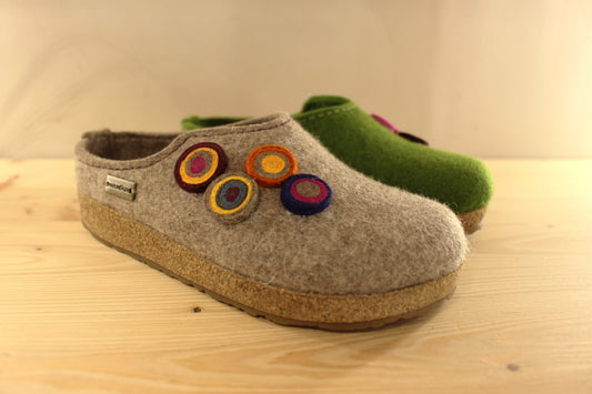  Chaussons HAFLINGER coupe sabot en pure laine de couleur vert ou beige. Déco de boutons colorés en laine sur le côté du coup de pied. Découpe asymétrique pour un bon maintien. 