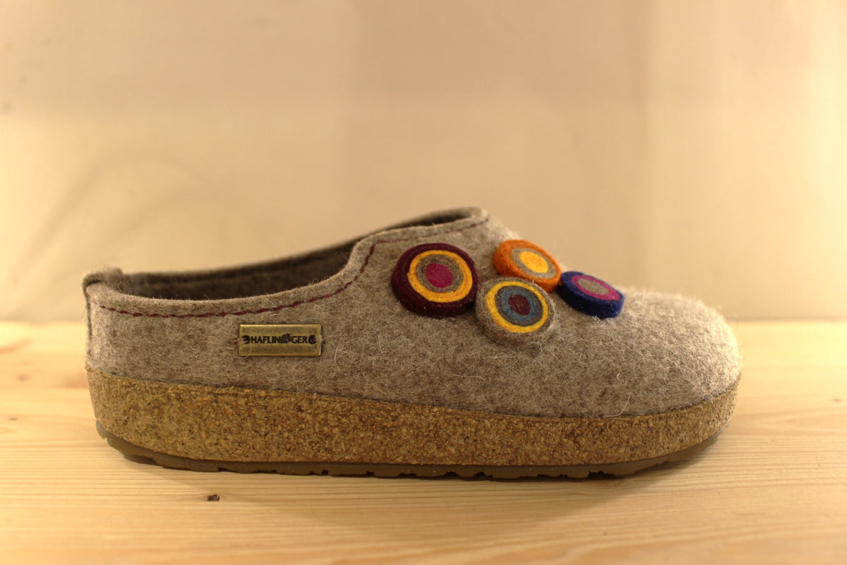  Chaussons HAFLINGER coupe sabot en pure laine de couleur vert ou beige. Déco de boutons colorés en laine sur le côté du coup de pied. Découpe asymétrique pour un bon maintien. 