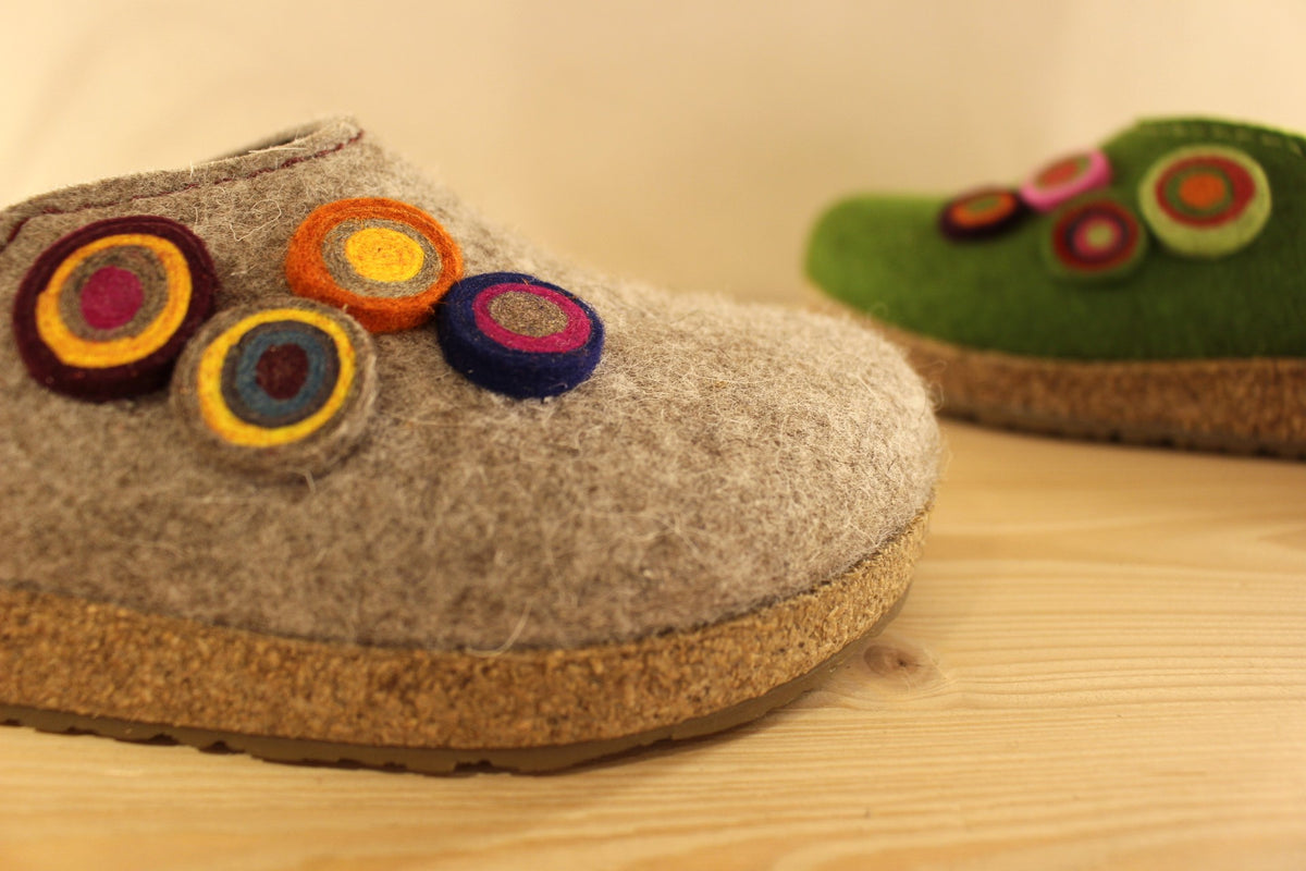  Chaussons HAFLINGER coupe sabot en pure laine de couleur vert ou beige. Déco de boutons colorés en laine sur le côté du coup de pied. Découpe asymétrique pour un bon maintien. 