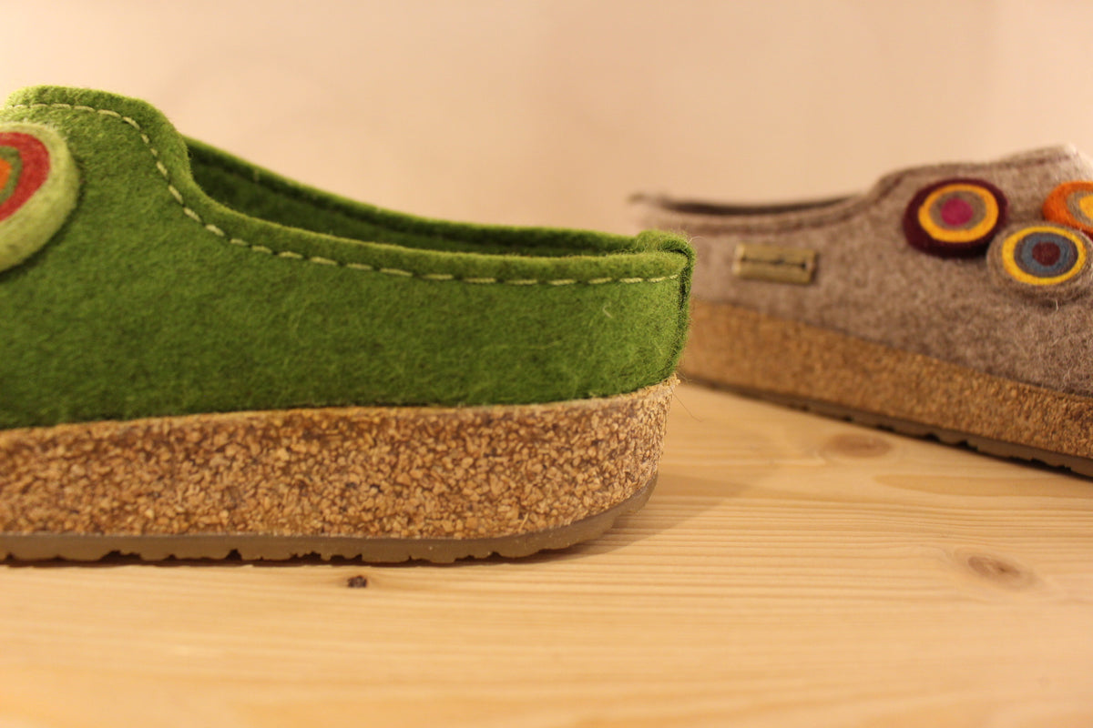  Chaussons HAFLINGER coupe sabot en pure laine de couleur vert ou beige. Déco de boutons colorés en laine sur le côté du coup de pied. Découpe asymétrique pour un bon maintien. 